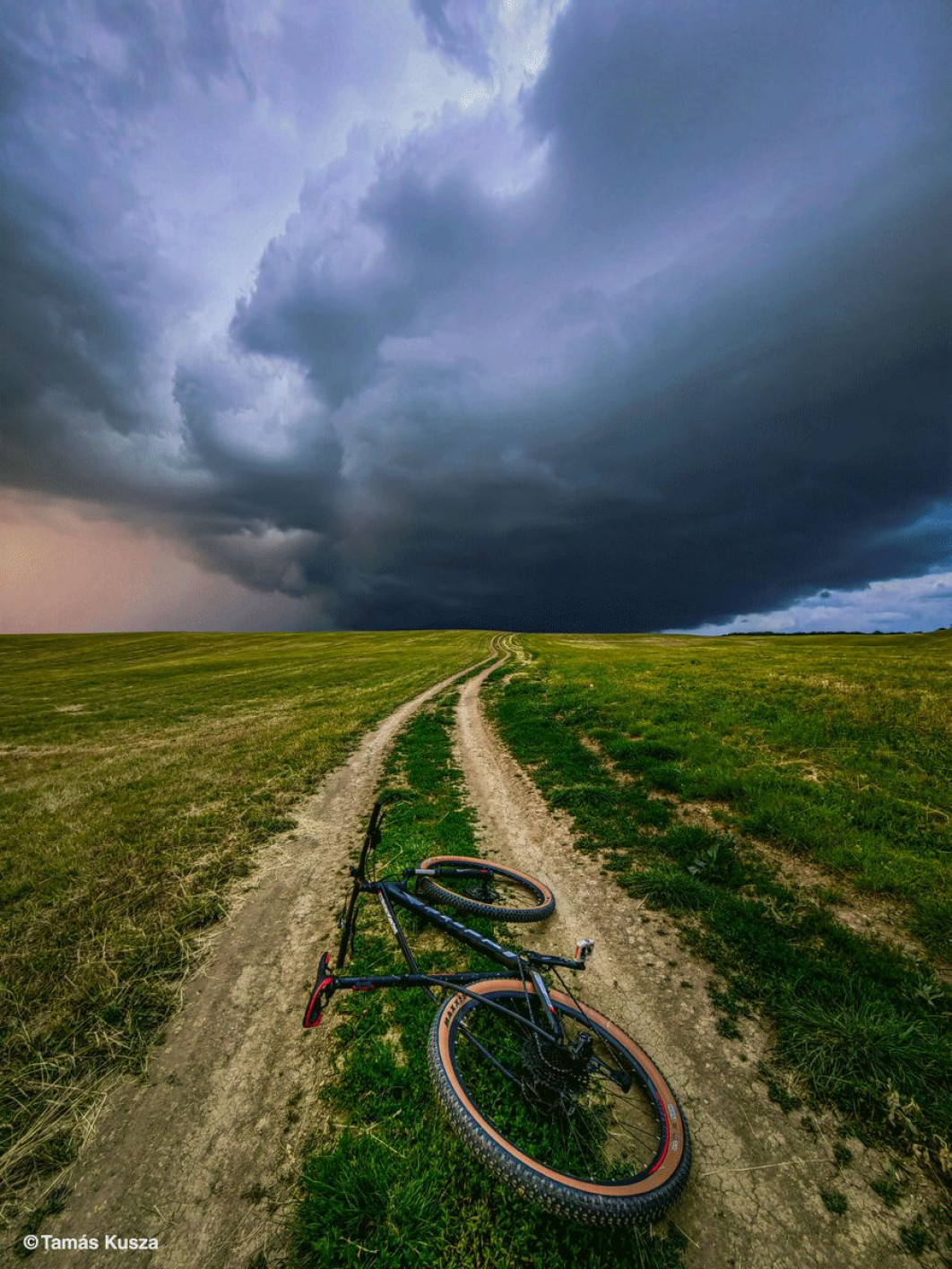 <strong>Wyróżnienie w kat. Fotografia mobilna,  fot. Tamás Kusza, Słowacja</strong><br /><br />

Polna droga przecina krajobraz w okolicach Ožďan na Słowacji, prowadząc wzrok w stronę nieba wypełnionego ciemnymi, wirującymi chmurami. Pośrodku ścieżki leży rower górski, a jego właściciel zatrzymał się, by przyjrzeć się nadchodzącej burzy. „Zawsze fascynowały mnie burze” - mówi fotograf Tamás Kusza. „Uwielbiam patrzeć, jak niebo ciemnieje, chmury schodzą coraz niżej, a majestatyczna siła natury powoli się ujawnia. Gdy tylko mogę, wsiadam na rower i jadę tam, gdzie ziemia spotyka się z niebem - tam, gdzie rodzą się burze.”<br /><br />

Tego dnia na horyzoncie zaczęły gromadzić się ciężkie chmury. „Wiedziałem, że nadchodzi wyjątkowy moment. Jechałem dalej polną drogą, aż musiałem się zatrzymać  widok był niemal paraliżujący. Odstawiłem rower, wyjąłem aparat i stanąłem naprzeciw serca burzy. To była chwila przed ciszą - moment, w którym spotkały się potęga natury i moja własna odwaga. Świat zamarł. Czy zostanę, by sfotografować burzę, czy zawrócę? Wiedziałem jedno: zawsze zmierzam w jej kierunku.”<br /><br />

Letnie burze, takie jak ta, są w Europie Środkowej zjawiskiem powszechnym - często przynoszą ulewne deszcze, silny wiatr i błyskawice. Dla Kuszy to właśnie ich nieprzewidywalność stanowi największy magnes, który każe mu podążać w stronę horyzontu. Wraz z postępującymi zmianami klimatycznymi region ten doświadcza coraz dłuższych, gorętszych lat, a pogoda staje się bardziej niestabilna - z częstszymi gwałtownymi ulewami i szybszym rozwojem burz nad lądem.
