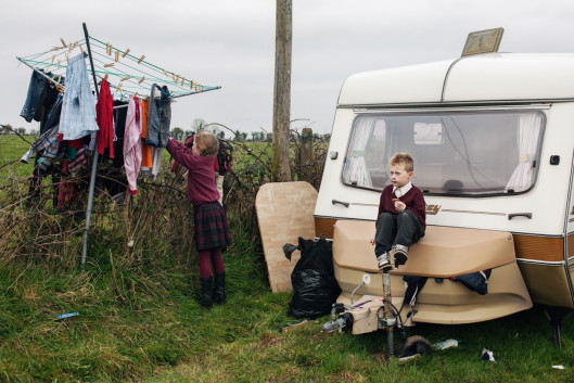 fot. Birte Kaufmann, „Travellersi”

Projekt „Travellersi” dokumentuje życie członków najliczniejszej mniejszości w Irlandii. Mowa o współczesnych nomadach wywodzących się z tradycji migracji zarobkowej. Dzisiaj cel tułaczki się zmienił, tzw. travellersi próbują na nowo odnaleźć swoją tożsamość w społeczeństwie Europy Zachodniej. Styl życia irlandzkich koczowników jest dosyć nietypowy, dlatego spotyka się z brakiem akceptacji ze strony reszty obywateli. Ci ludzie żyją jakby w równoległej rzeczywistości, która rządzi się swoimi prawami, i w której obowiązuje tradycyjny podział ról. Osoby z zewnątrz rzadko mają wstęp do tego hermetycznego świata.

Po dziś dzień niektóre rodziny rozbijają nielegalne obozowiska przy drodze. Żyją bez prądu, bieżącej wody, ani sanitariatów, choć władze zapewniają im specjalne pola kempingowe, gdzie mogą parkować swoje wozy.

Pierwszy raz do Irlandii wybrałam się VW „ogórkiem” w 2011 roku. Na zdjęciach chciałam uchwycić styl życia travellersów i wartości, którymi się kierują. Nie zamierzałam idealizować ich zwyczajów. Chciałam pokazać świat współczesnych nomadów, którzy polują na króliki, nie mogą się obejść bez koni, od dziecka zmagają się z trudami życia i walczą z nudą.

Podczas mojego pierwszego spotkania z irlandzkimi tułaczami poznałam pewną liczną rodzinę. W miarę upływu czasu zdobywałam ich zaufanie. W końcu zgodzili się, żebym z nimi zamieszkała. Ja i mój aparat staliśmy się częścią ich codziennego życia